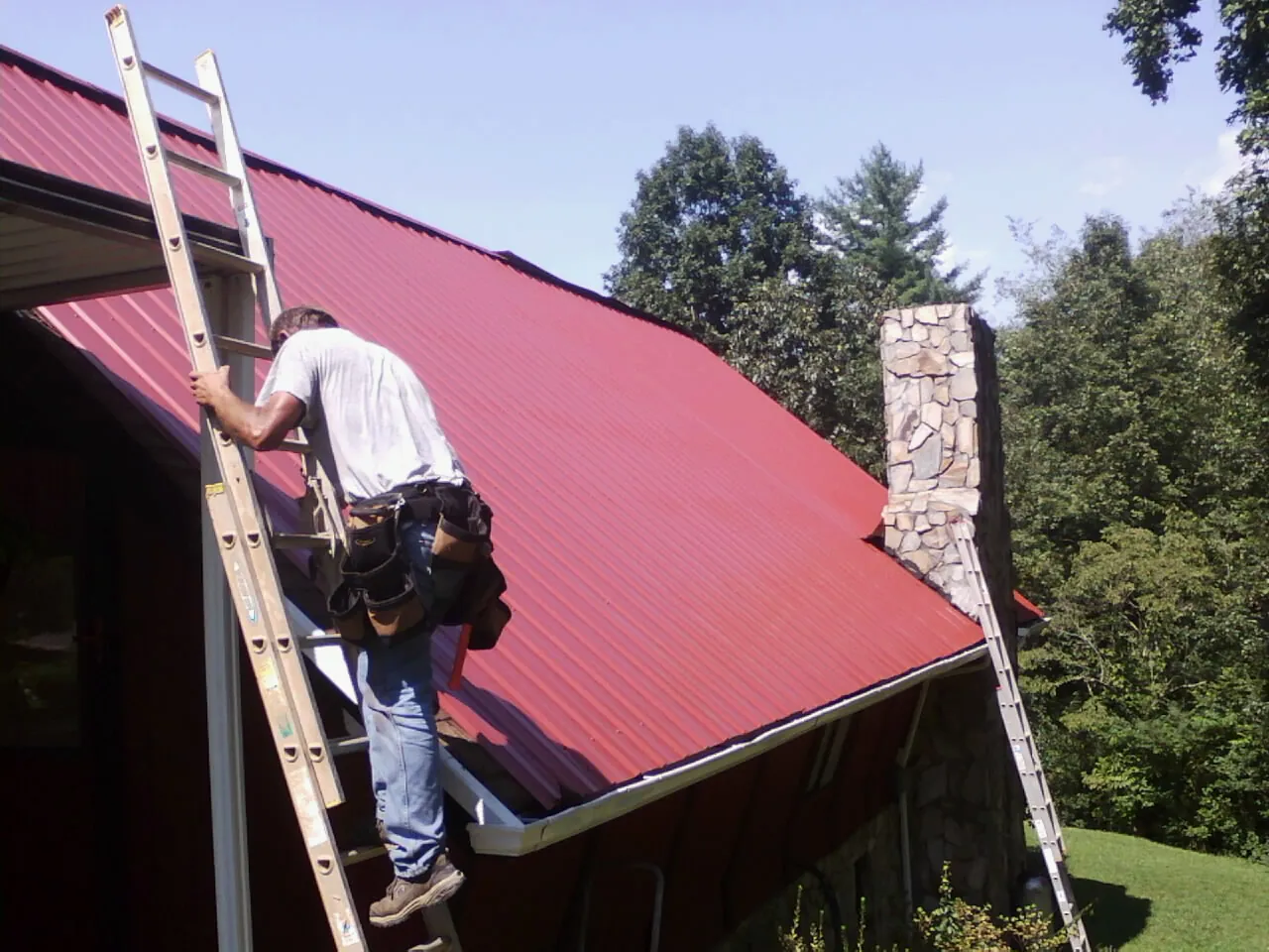 Roofer on a ladder beside a red metal roof and stone chimney — Soffit & Fascia Repair by Roofing Brothers in Leakey